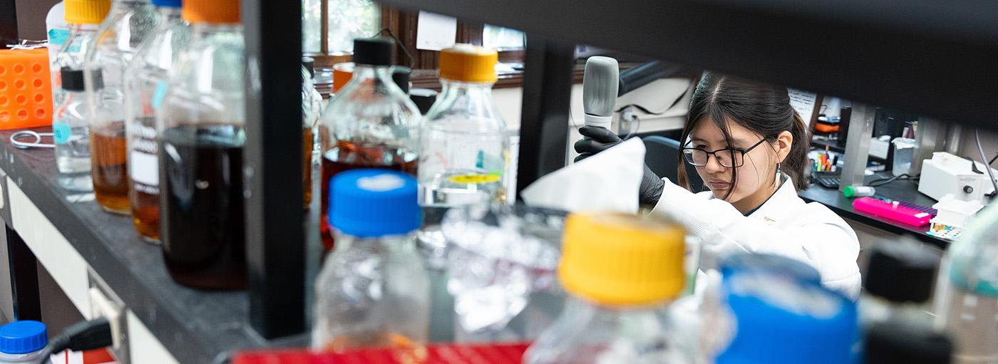 一名妇女站在摆满彩色瓶子的实验室架子后面，用移液管测量液体.